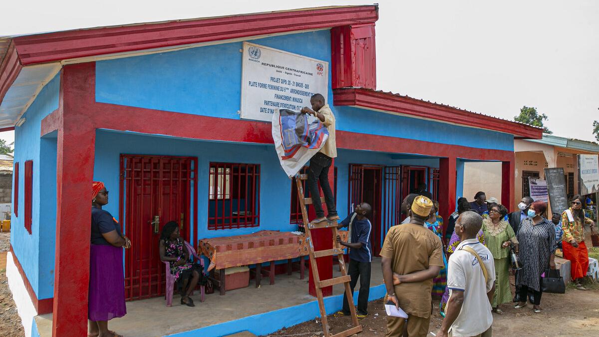 A man on a ladder unveils a sign on a new single-story building in front of a crowd