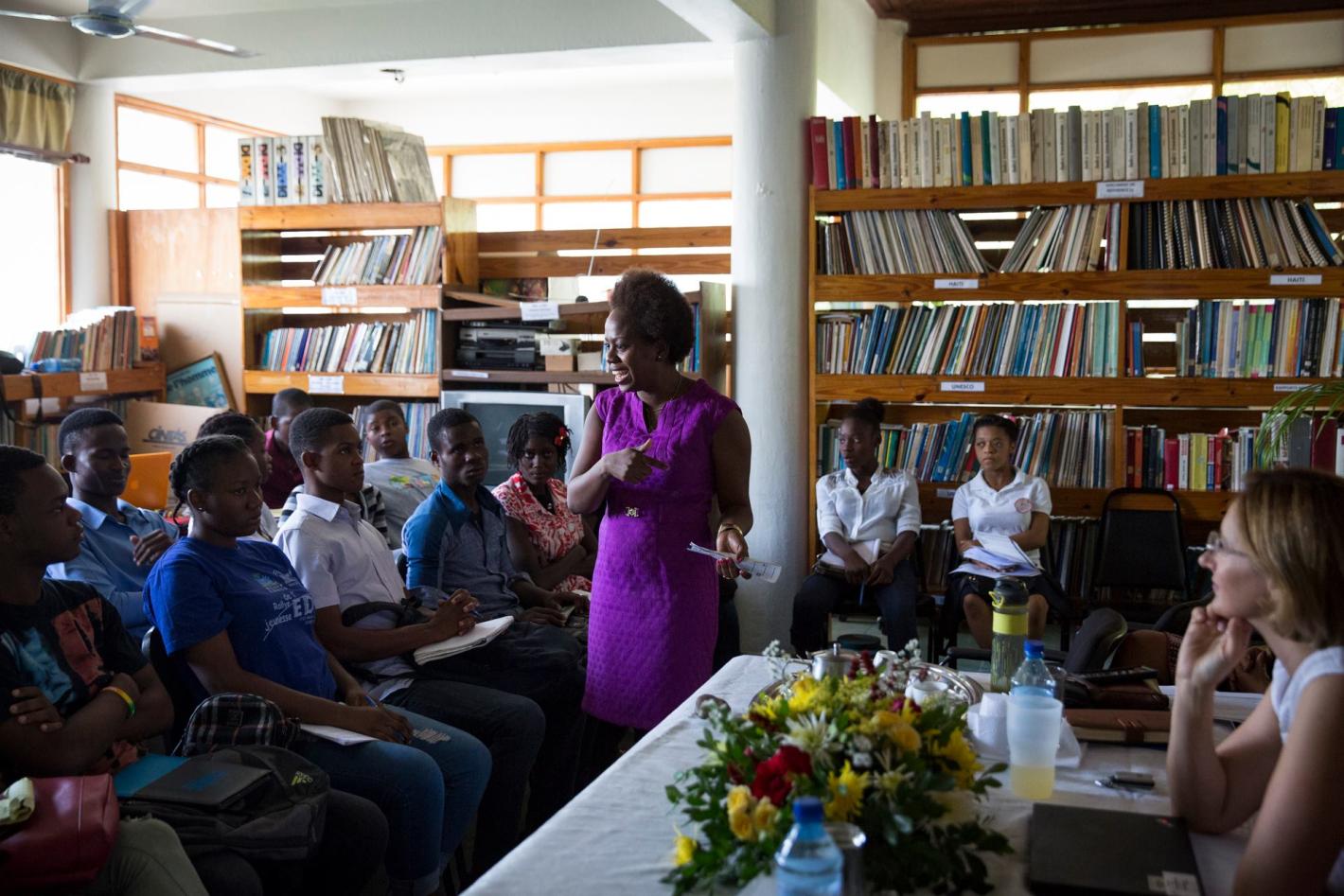 A group of people sitting in a library listening to a woman in a purple dress speaking to the group.